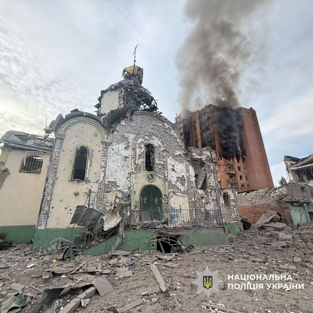 Зруйнований російським обстрілом храм у Краматорську: пошкоджені стіни, вікна, дах і купол, навколо лежать уламки. Позаду храму горить пошкоджена багатоповерхівка, над нею здіймається густий дим.